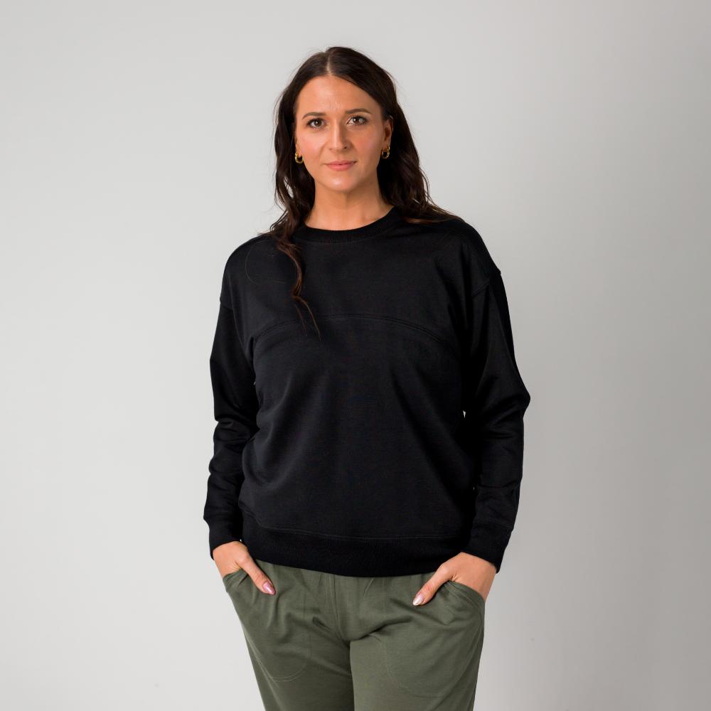 A woman modeling a black merino tencel blend crewneck nursing sweatshirt and olive green pants against a plain gray studio background