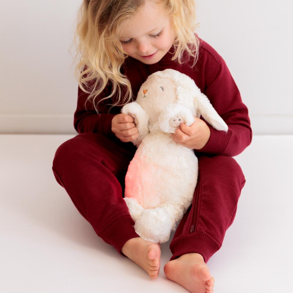 A young child with blonde hair sits on a white floor wearing a deep red merino fleece onesie, holding a fluffy white stuffed bunny with a pink belly.
