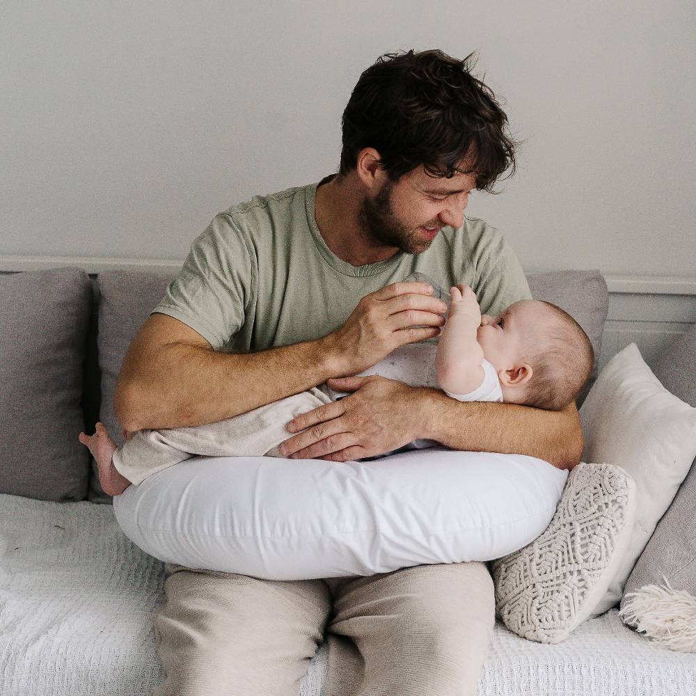 Man sitting on a couch holding a baby on a feeding pillow both wearing white pants.