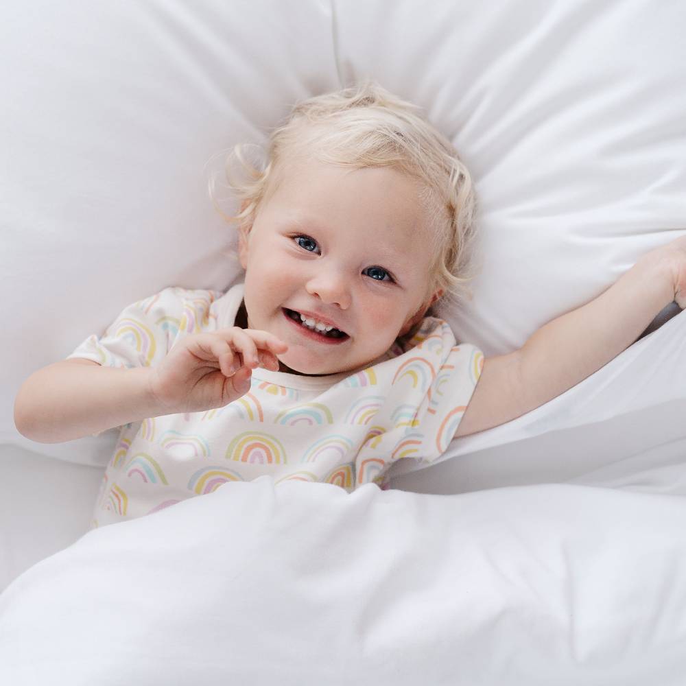 Child lying in bed with a white pillow and blanket, wearing pyjamas with rainbow patterns.