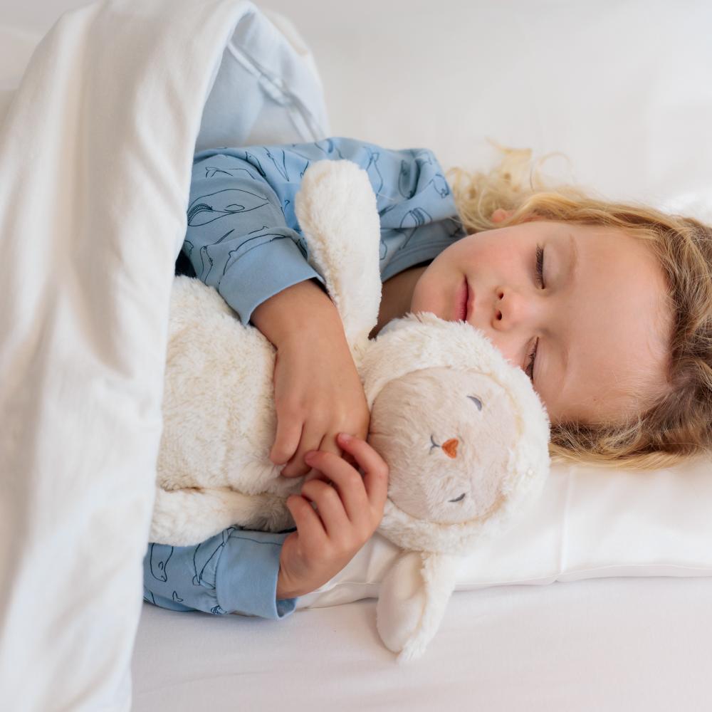 A young child sleeping soundly under a white blanket, hugging a plush white stuffed sheep nightlight and sound machine with a soft beige face and small orange nose.