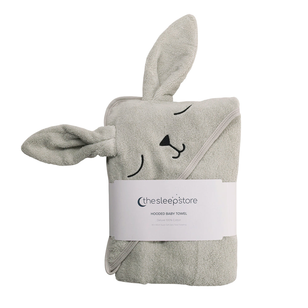 A folded cotton terry hooded baby towel in a light gray color, featuring a bunny hood with embroidered eyes, nose, and closed-eyes expression. A white paper band with the brand 'thesleepstore' is wrapped around the middle, set against a plain white background.