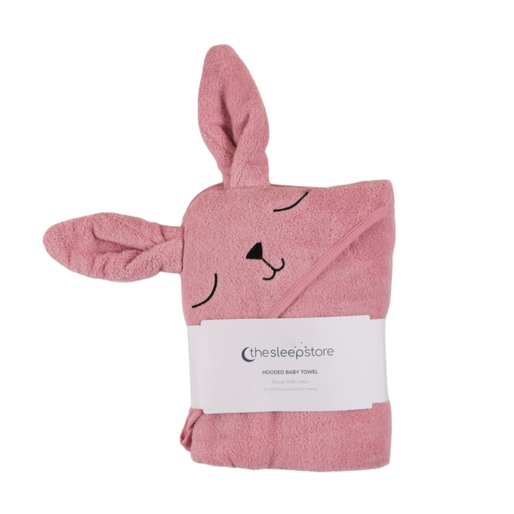 A pink cotton terry hooded baby towel shaped like a bunny, with embroidered closed eyes, a black nose, and ears on the sides, wrapped in a white paper band labeled 'thesleepstore'.