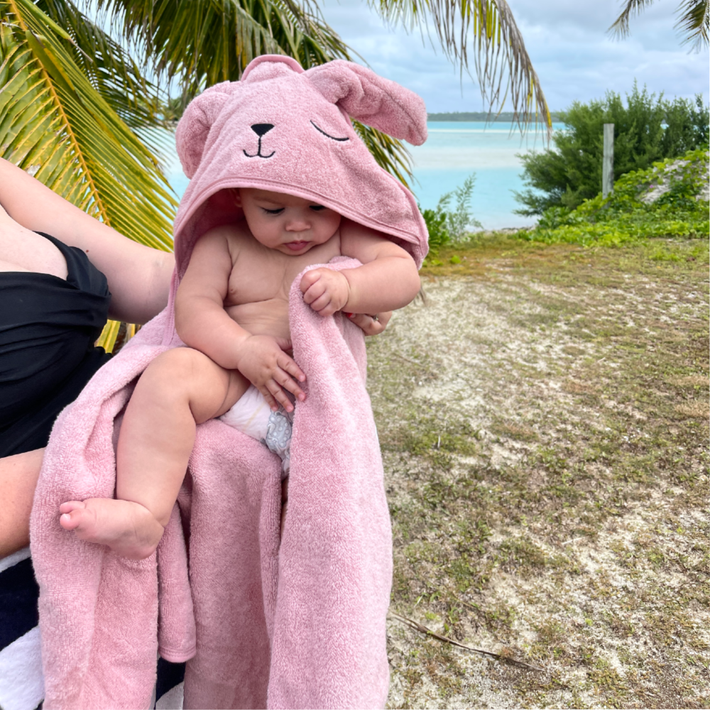 A baby wrapped in a pink bunny-shaped hooded towel outdoors near palm trees and water, with part of an adult holding the child visible on the left.
