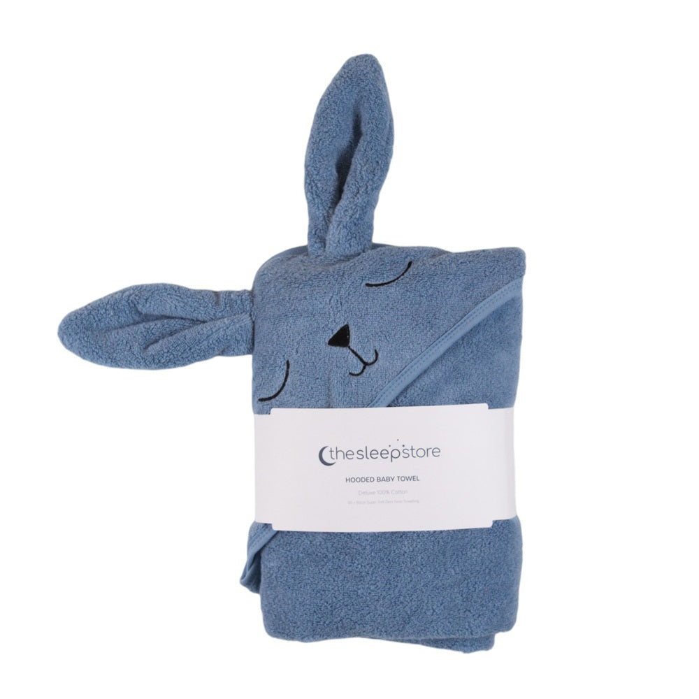 A folded blue hooded terry cotton baby towel shaped like a bunny, with embroidered closed eyes and nose, long ears, and a white paper band reading 'thesleepstore' around the center, on a plain white background.