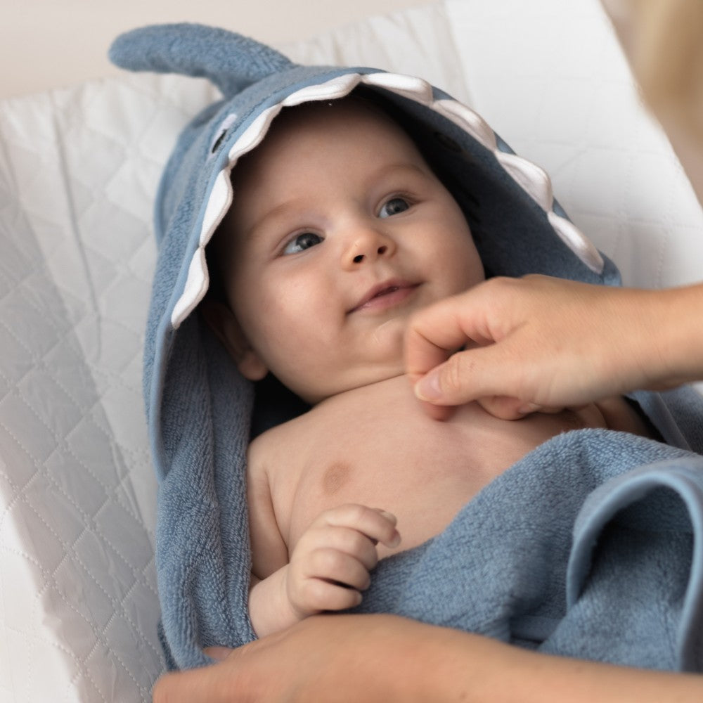 A baby wrapped in a blue whale-shaped hooded towel, lying on a white changing pad while an adult hand gently touches the baby's chest.