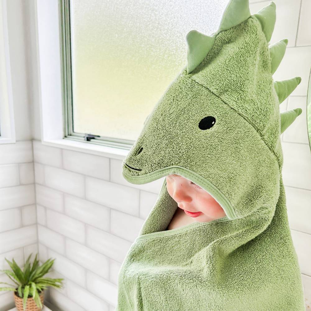 A baby wrapped in a light green dinosaur-themed hooded cotton terry towel, peeking out from the side of the hood with a black embroidered eye, set in a bright bathroom next to a window.