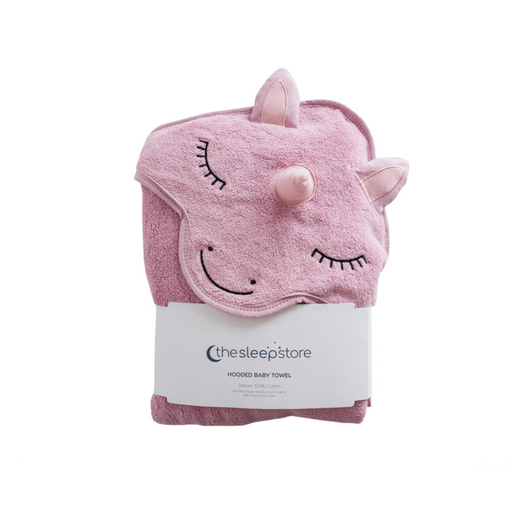 A pink hooded cotton terry baby towel shaped like a unicorn head, folded and wrapped in a white paper band labeled 'thesleepstore', on a plain white background.