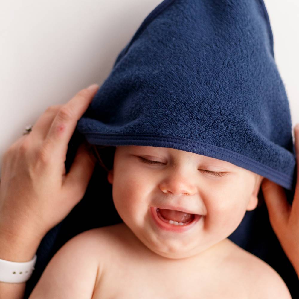A close-up of a baby wearing a navy blue cotton terry hooded towel, with an adult's hands adjusting the hood. The baby is smiling with eyes closed against a plain light background.