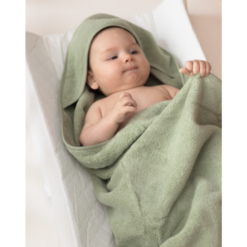 A baby wrapped in a soft sage green cotton terry hooded towel, lying on a white changing pad.