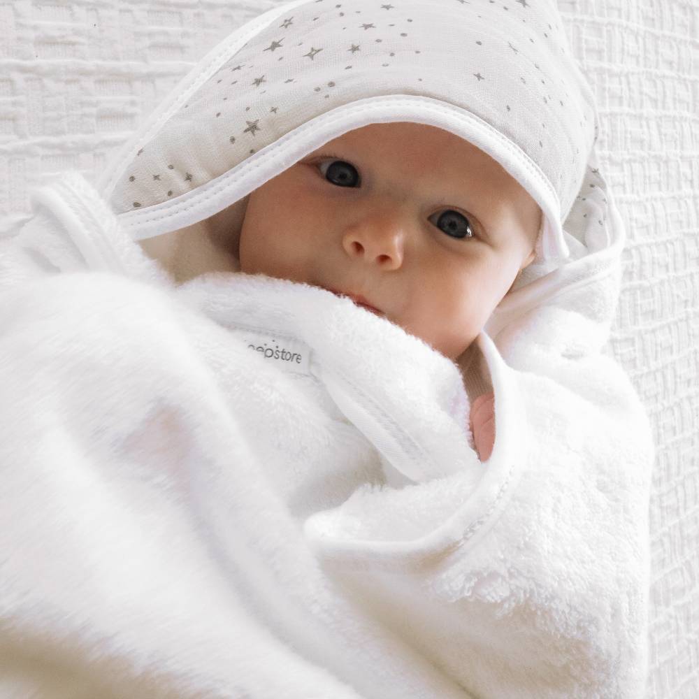 A close-up of a baby wrapped in a white plush towel hooded wrap with a matching white hood decorated with small gray stars, lying on a textured white blanket. The brand name 'the sleep store' is visible on a small fabric tag.