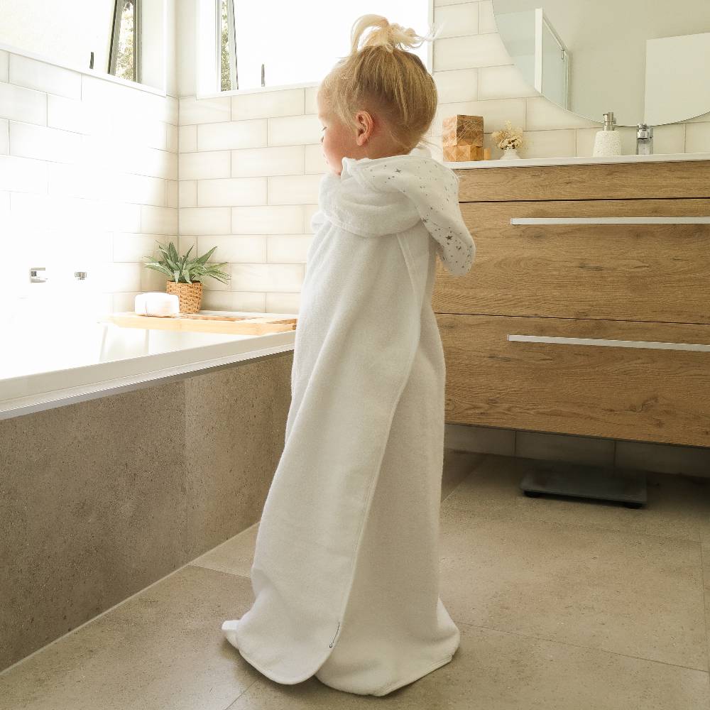 A young child with blonde hair in a high ponytail stands in a bathroom, wrapped in a soft white hooded towel . The bathroom features white subway tiles, a light wood vanity, and a bathtub.