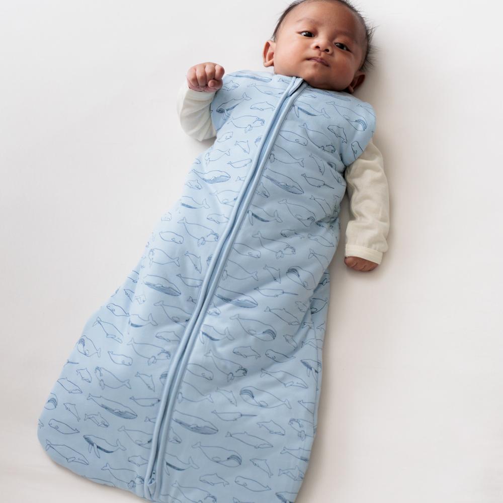 Baby lying on a white surface wearing a blue patterned sleeping bag.