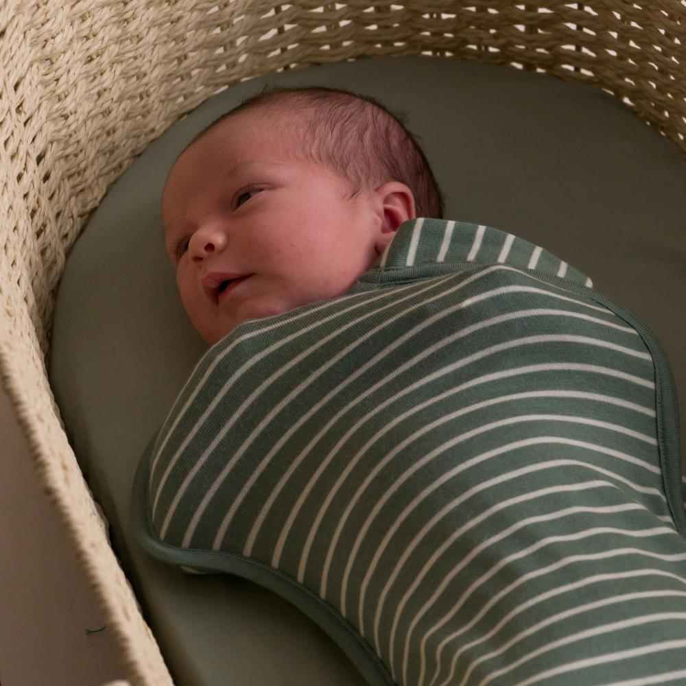 Newborn baby wrapped in a green and white striped merino swaddle inside a wicker basket.
