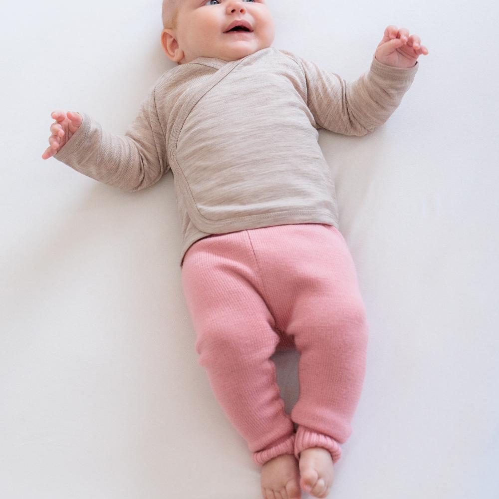 Baby lying on a white surface wearing a beige long-sleeve kimono style wrap cardigan and pink merino knitted pants.