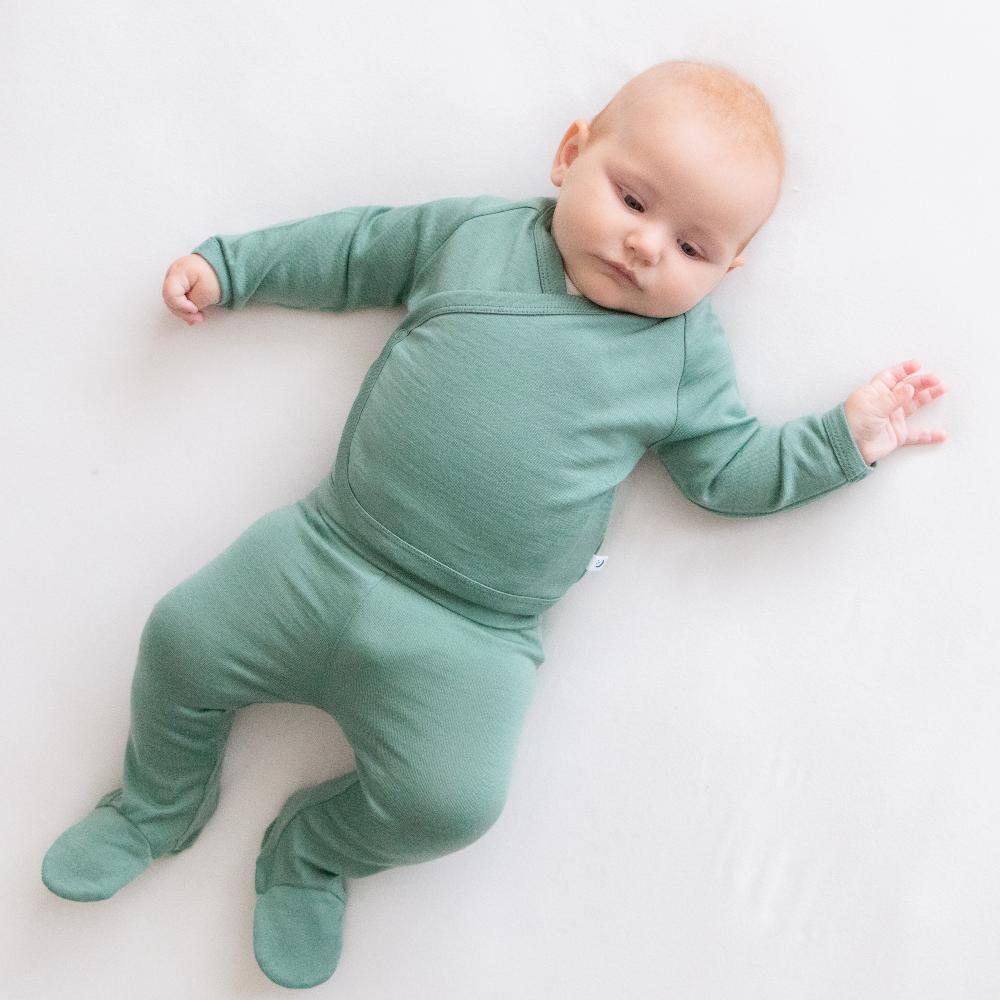 A baby lying on a white surface wearing a sage green long-sleeve jersey merino kimono cardigan and footed romper leggings