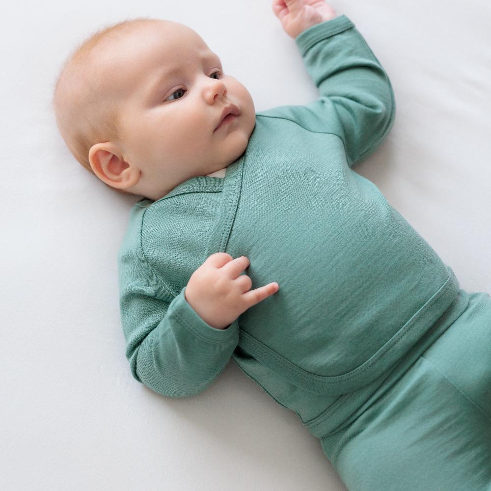A baby lying on a white blanket, wearing a sage green jersey merino kimono long-sleeve wrap cardigan and matching pants