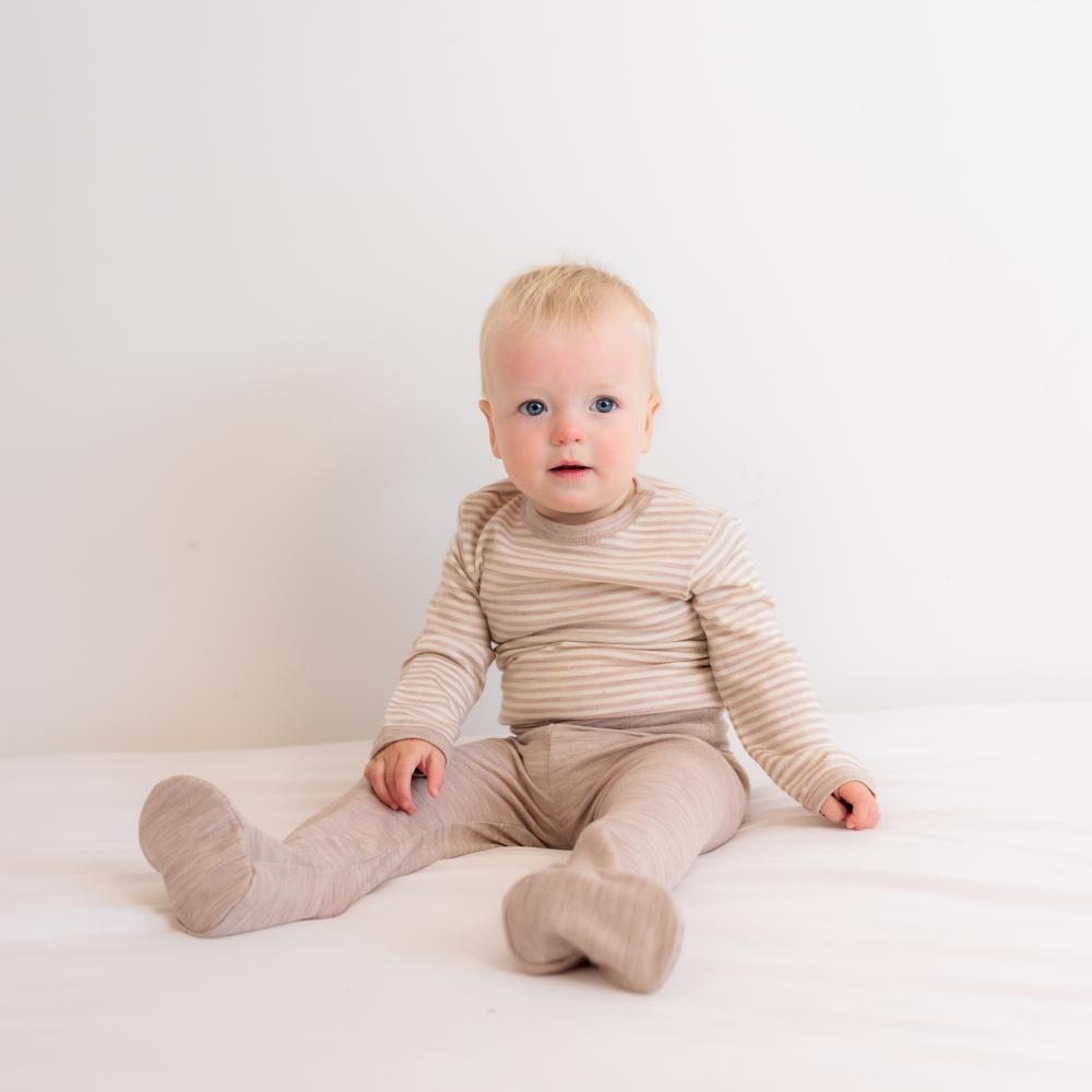 Baby sitting on a white surface wearing a beige striped jersey merino bodysuit and beige jersey merino footed leggings 