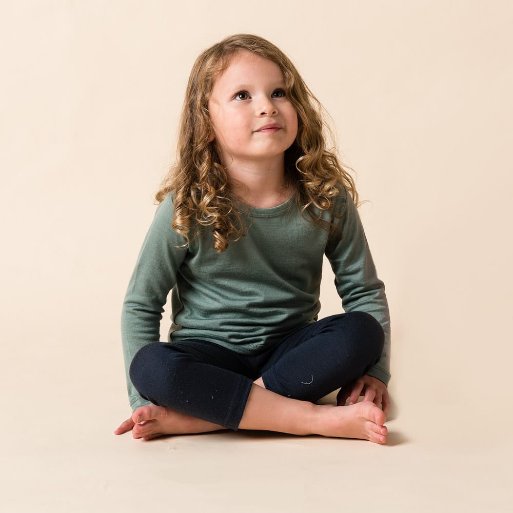 Young girl sitting cross-legged wearing a green merino long-sleeve shirt and dark pants on a beige background