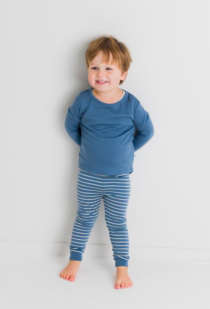 A toddler standing against a plain light gray wall wearing a jersey merino pyjama set including blue long-sleeve top and blue-and-white striped pants.