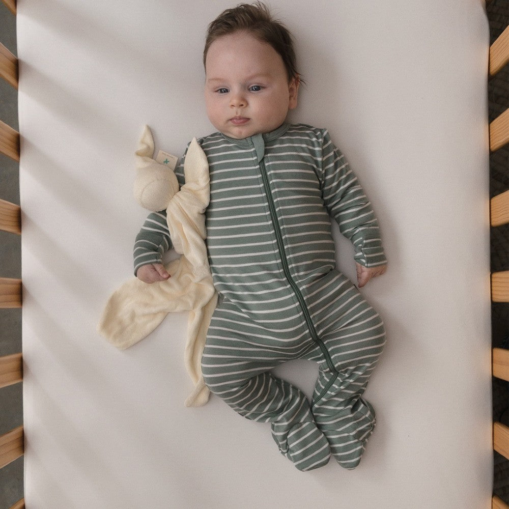 Baby in a green and white striped jersey merino onesie lying on a white blanket with a plush toy.