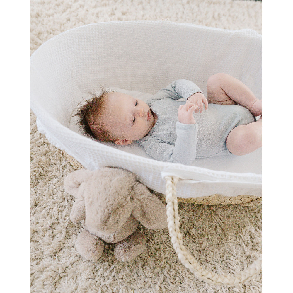A baby in a light blue long-sleeve bodysuit rests inside a white woven Moses basket on a beige shag rug, with a plush beige stuffed animal beside the basket.
