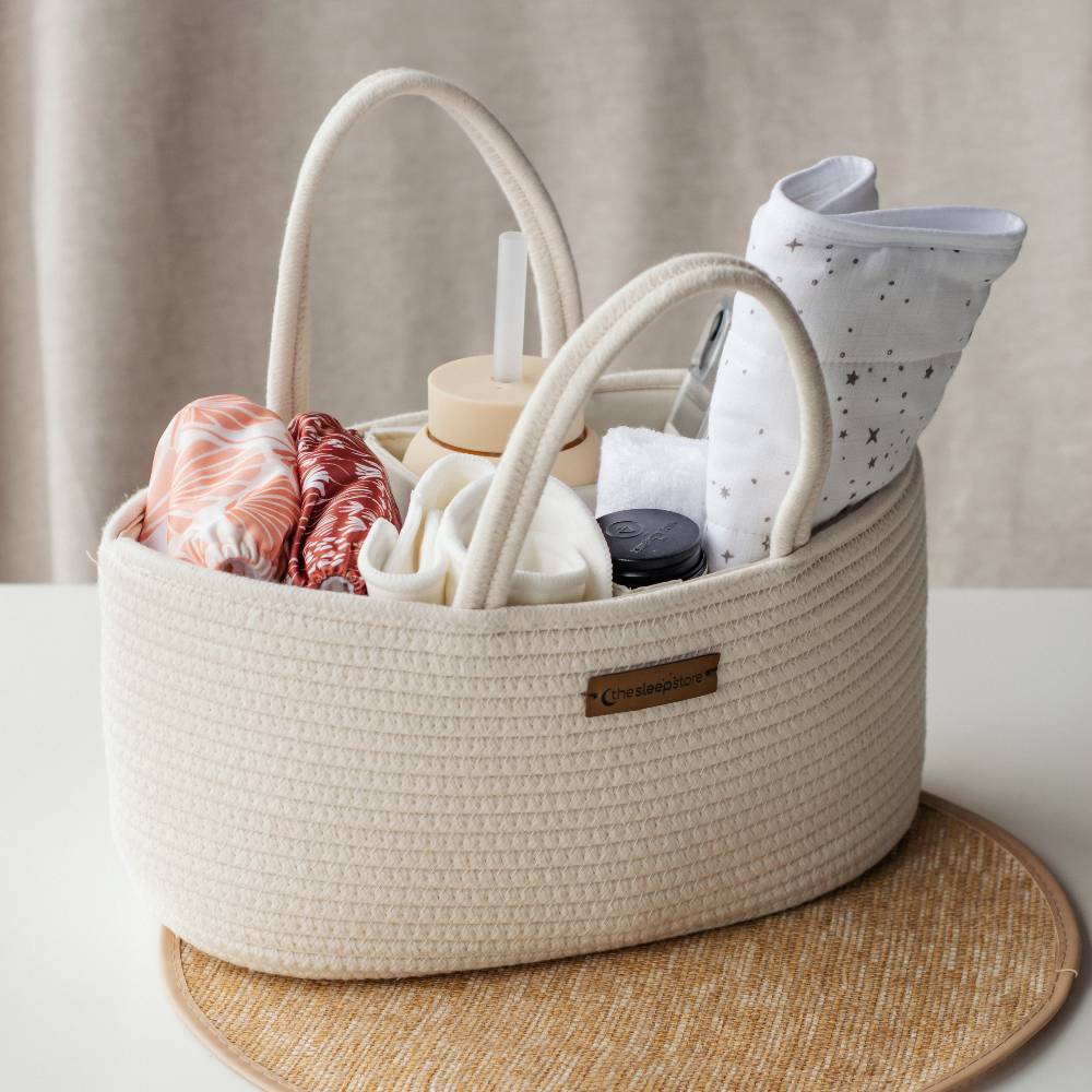 A cream-colored woven storage basket with dual top handles, filled with baby items including a patterned cloth, a white star-print cloth, a folded white towel, and a bottle. The basket sits on a round woven placemat on a white surface with a neutral beige curtain background.