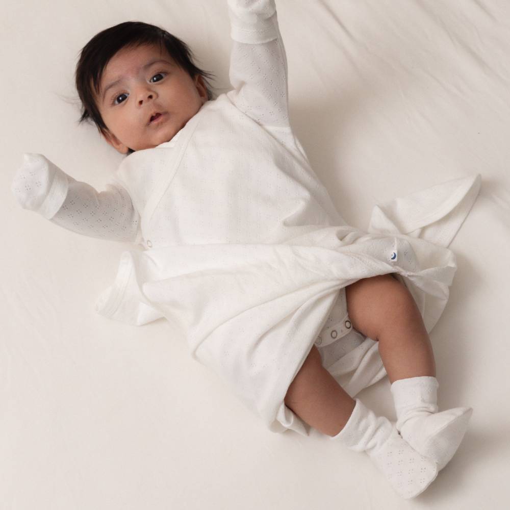 A baby lying on a white blanket, wearing a white long-sleeve sleep gown and white pointelle socks.