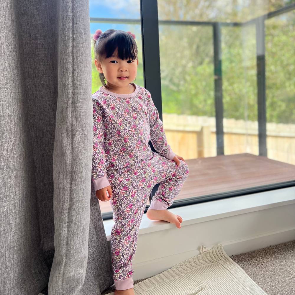 Child in cotton purple floral pajamas standing by a window with a view of greenery