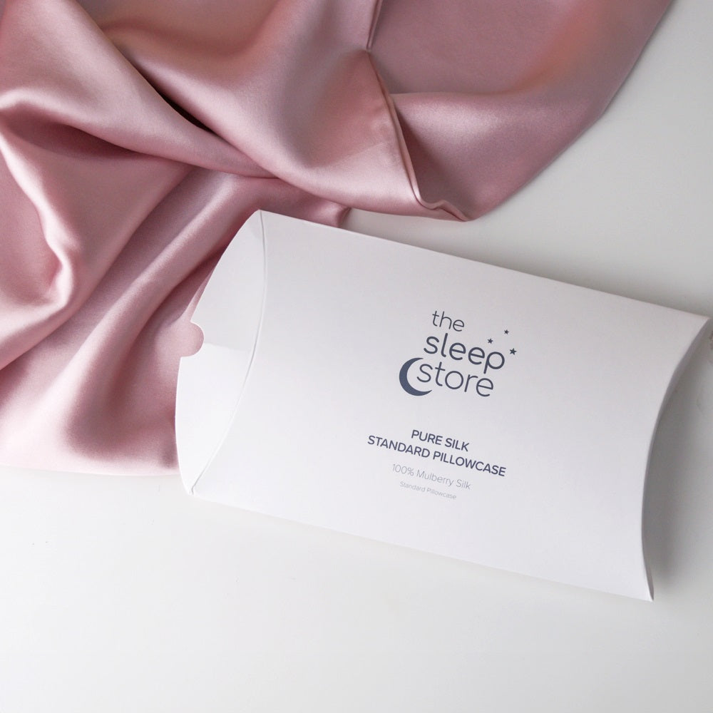 A white pillowcase packaging box with the brand name 'the sleep store' printed on it, styled on a white surface with draped pink satin fabric.