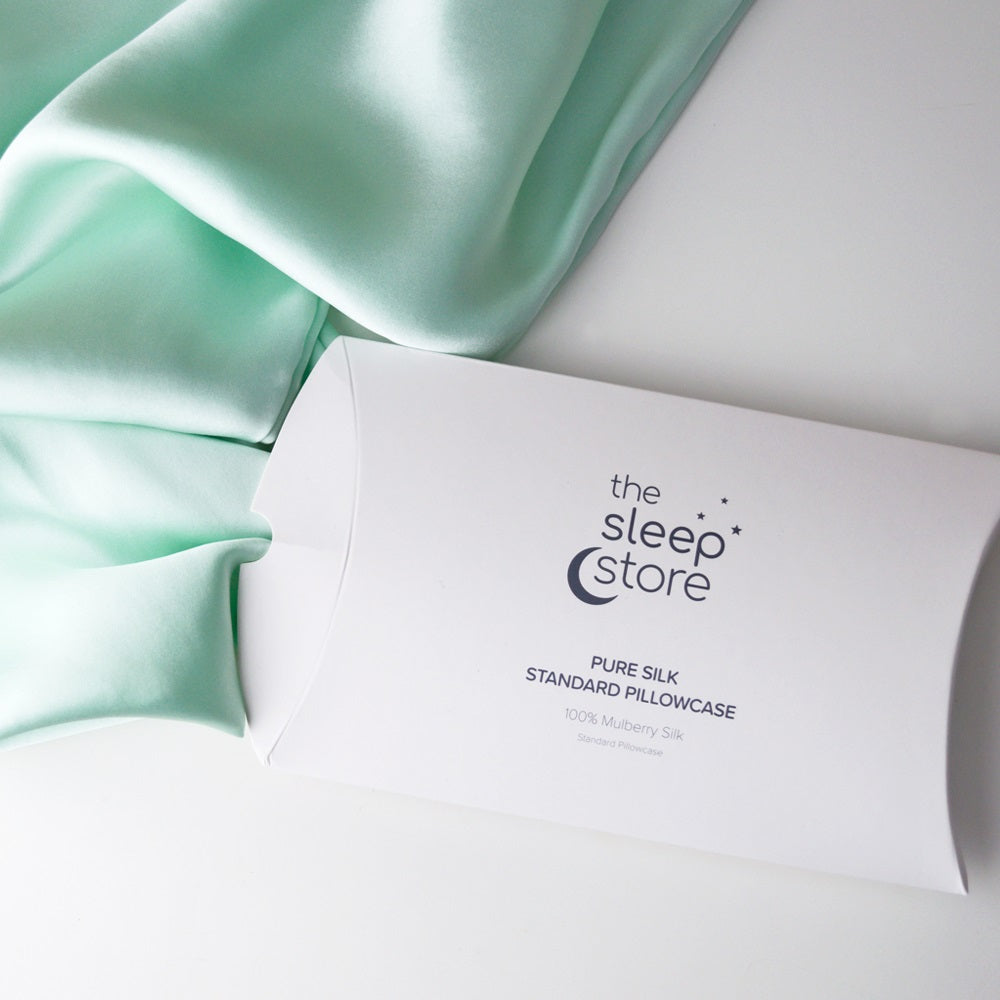 A white pillowcase box printed with 'the sleep store' and 'PURE SILK STANDARD PILLOWCASE' lies on a white surface next to draped mint green silk fabric.