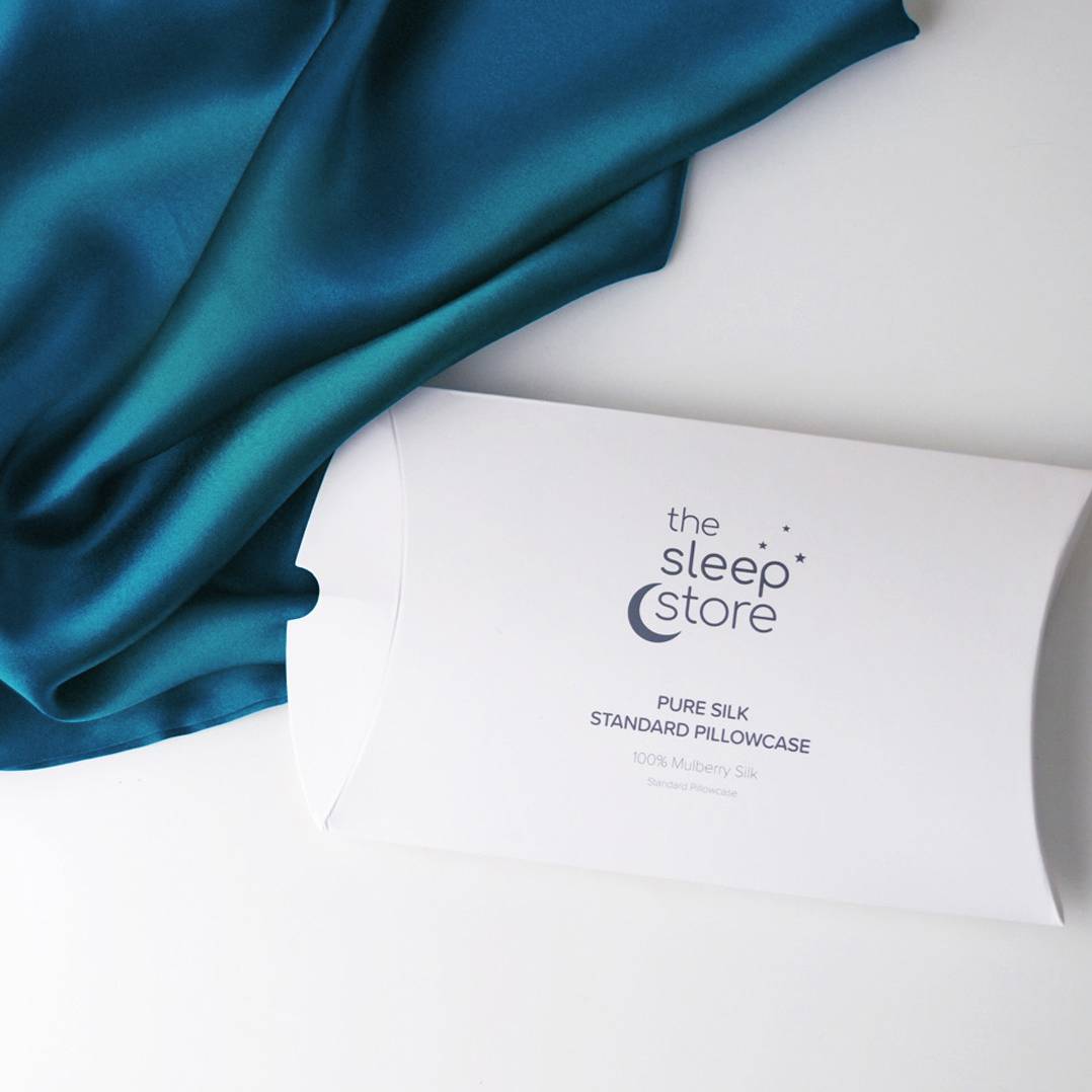 A white pillowcase packaging box printed with 'the sleep store' and 'PURE SILK STANDARD PILLOWCASE' lies on a white surface next to draped teal satin fabric.