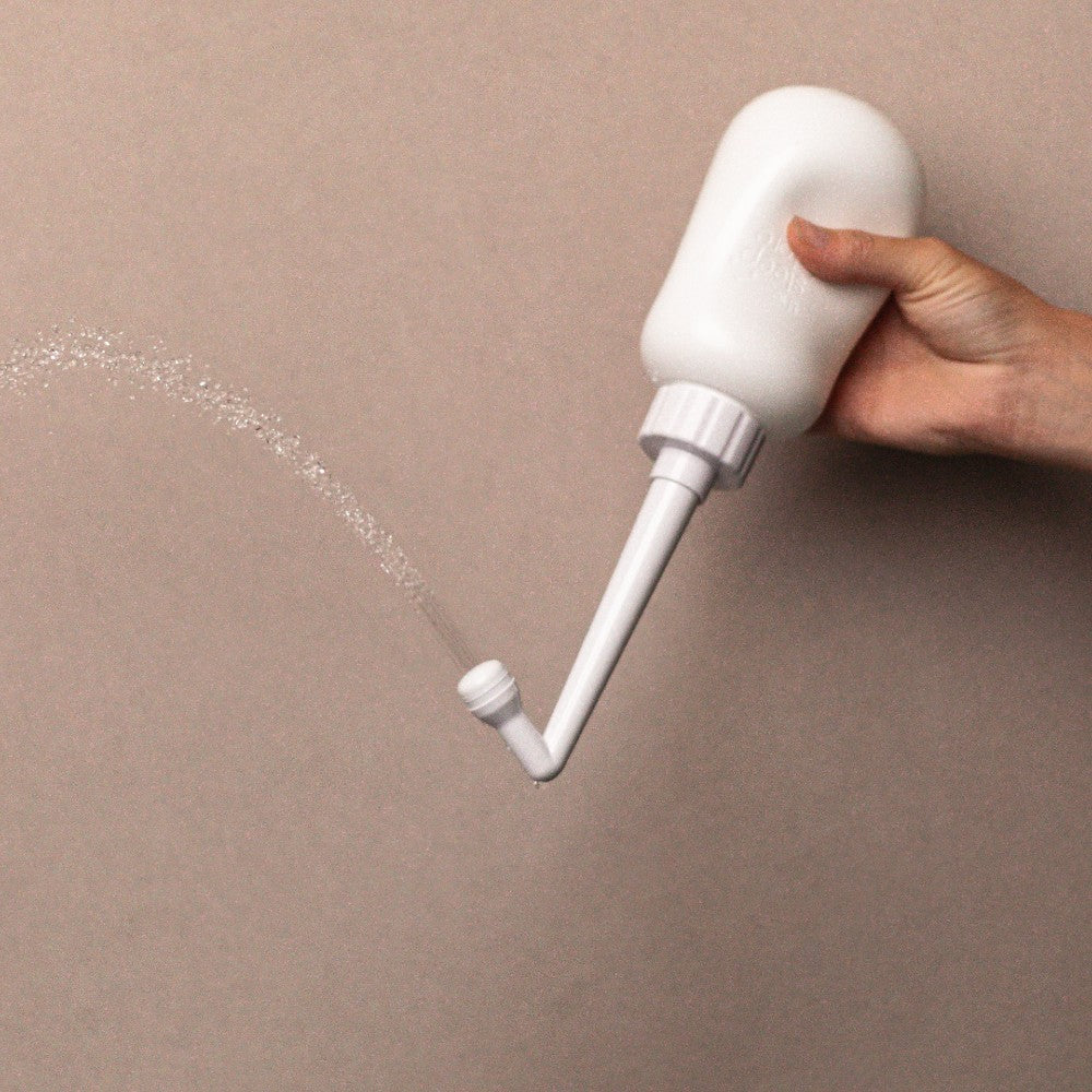 White peri bottle with a nozzle being held, releasing a stream of water on a beige background