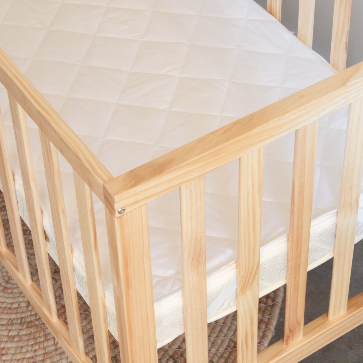 Wooden crib with white mattress and beige sheet and quilted wool protector on a dark background