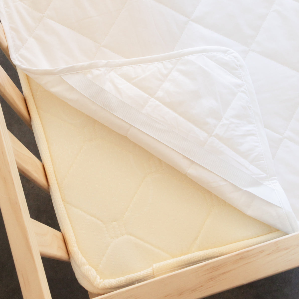 Wooden crib with white mattress and beige sheet and quilted wool protector on a dark background