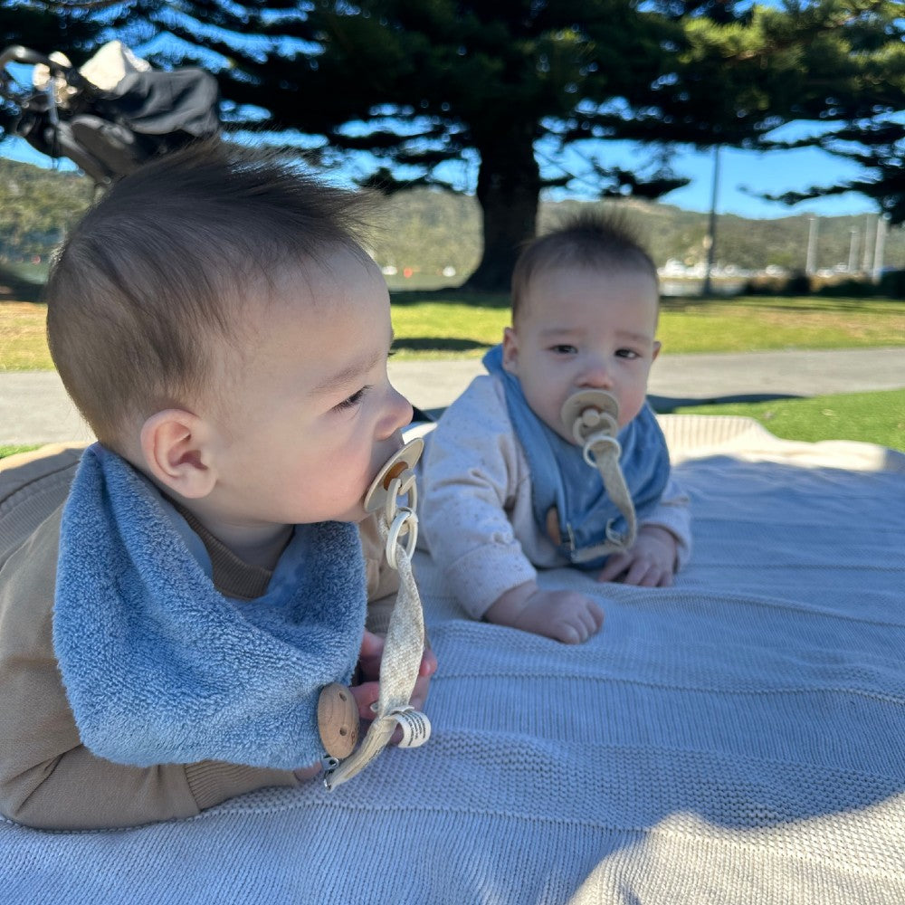 Two babies with blue bibs sitting on a blanket outdoors.