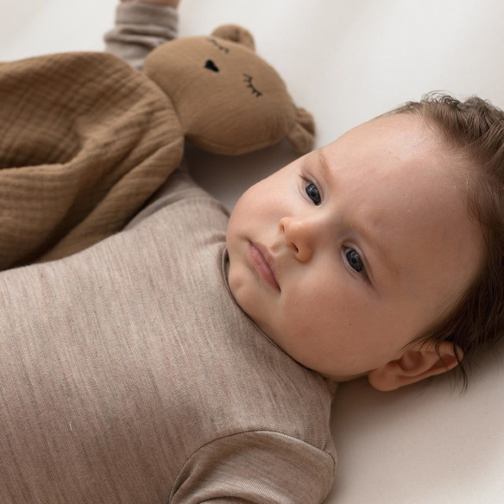 Baby lying on a white surface with a brown teddy bear muslin comforter next to them wearing a brown merino long sleeve bodysuit 