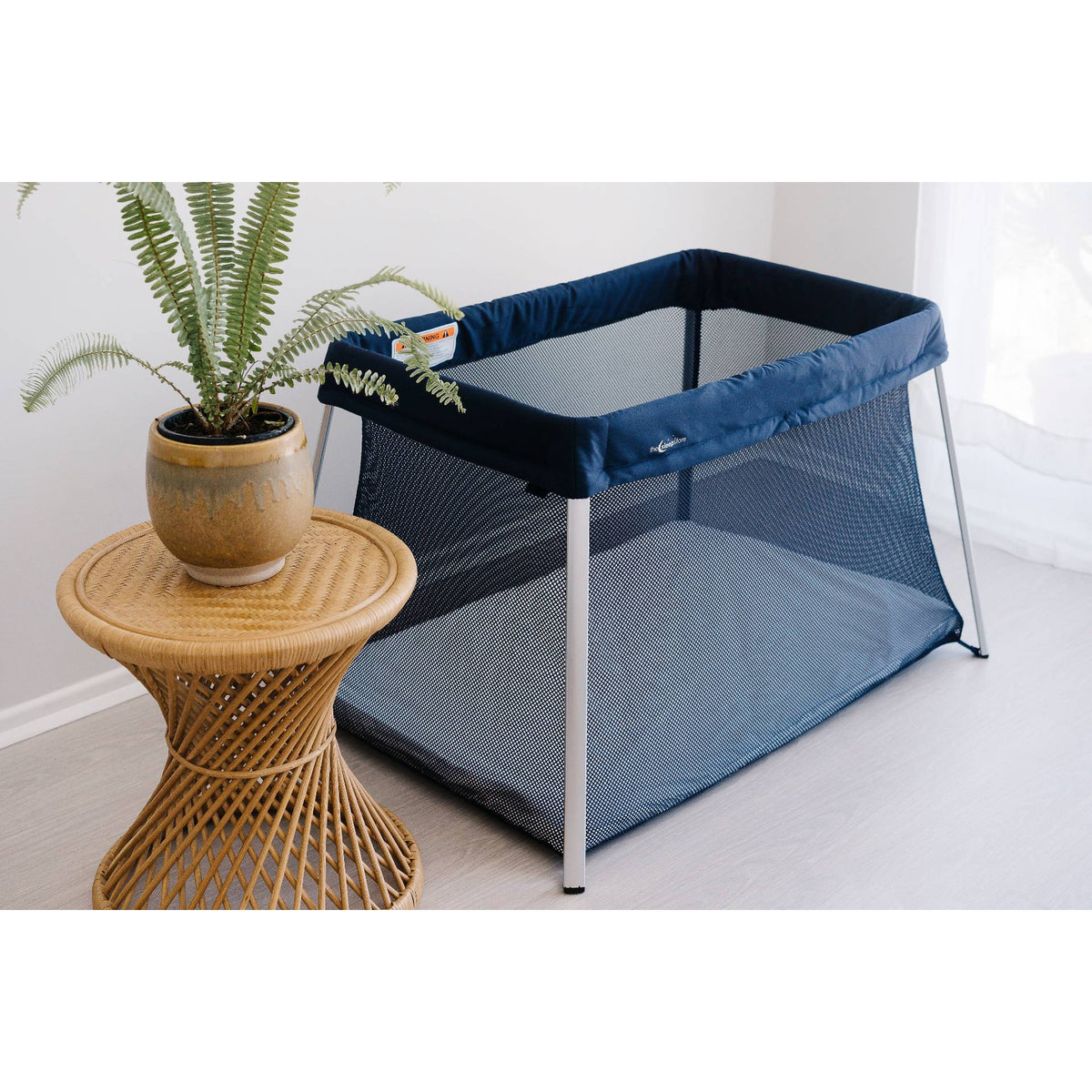 Portable travel cot next to a small table with a plant on a light-colored floor.