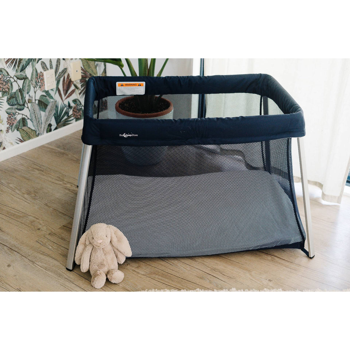 Portable baby travel cot with a plush toy on a wooden floor in a room with plants and a plant wall.