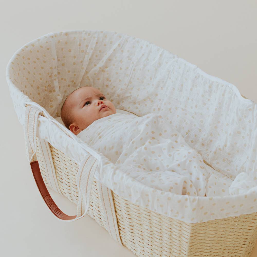 Baby wrapped in a white muslin blanket with spots lying in a wicker Moses basket.