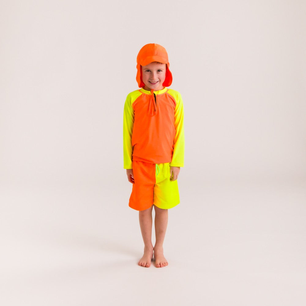 Child wearing a bright orange and yellow rash top and board shorts with a hat on a light gray background