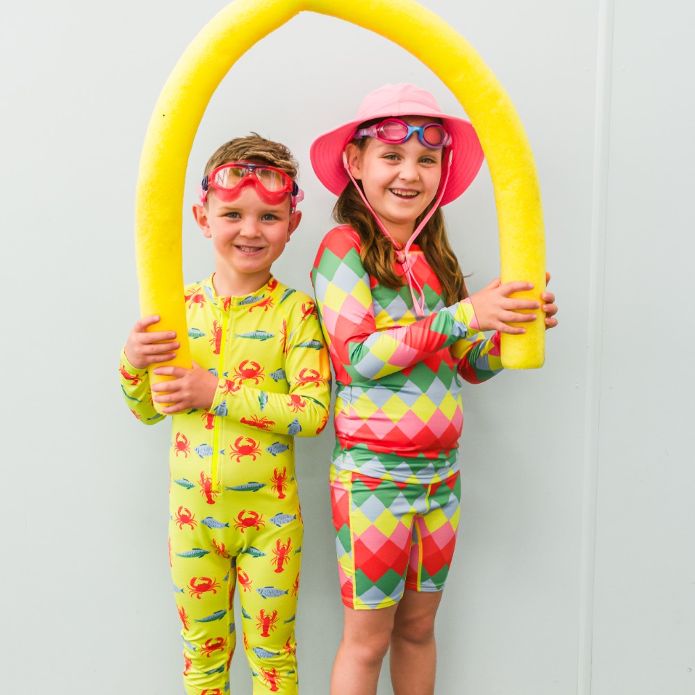 Two children in colorful swimwear holding a yellow pool noodle against a white background