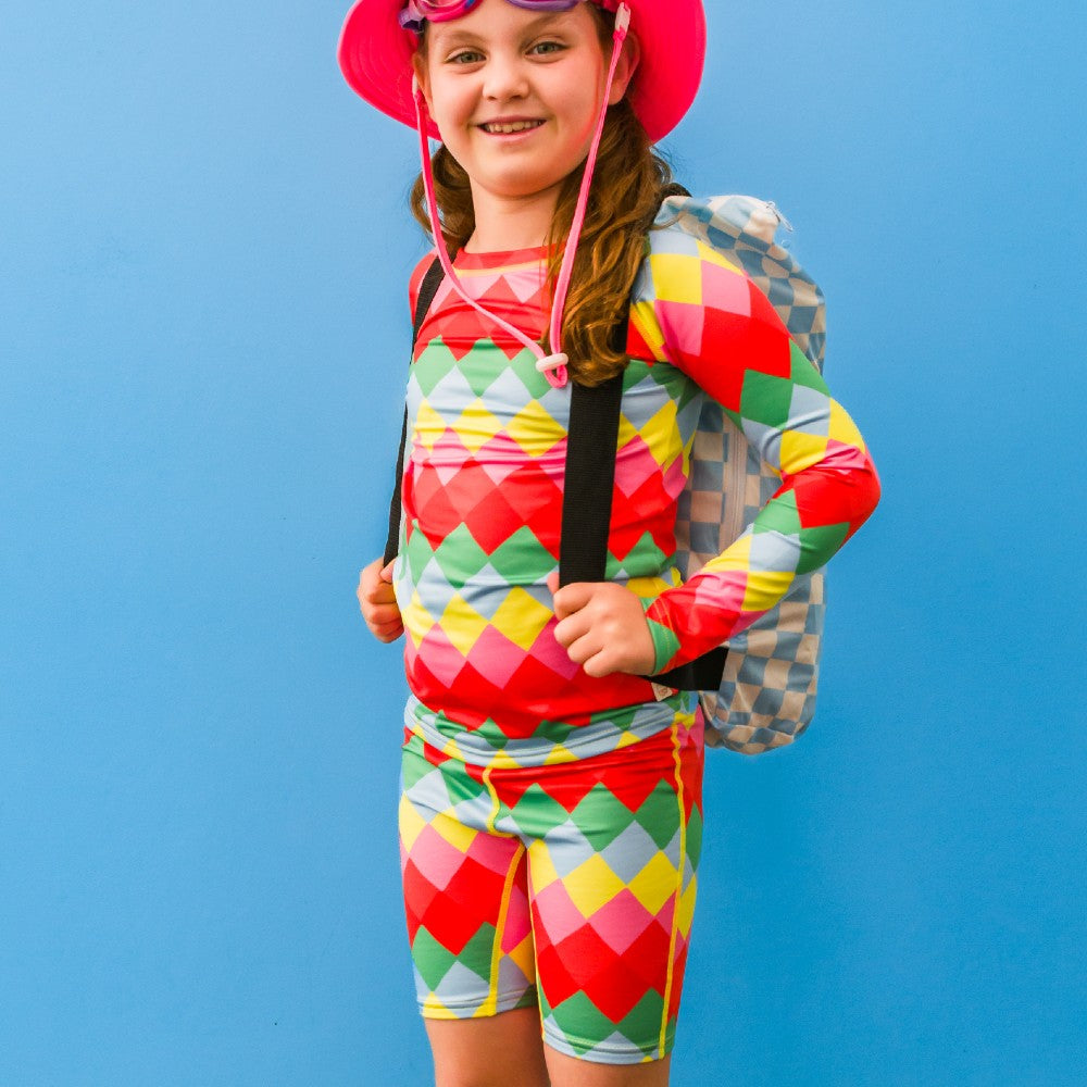 Child wearing a colorful diamond-patterned rash top and shorts with a pink hat against a blue background