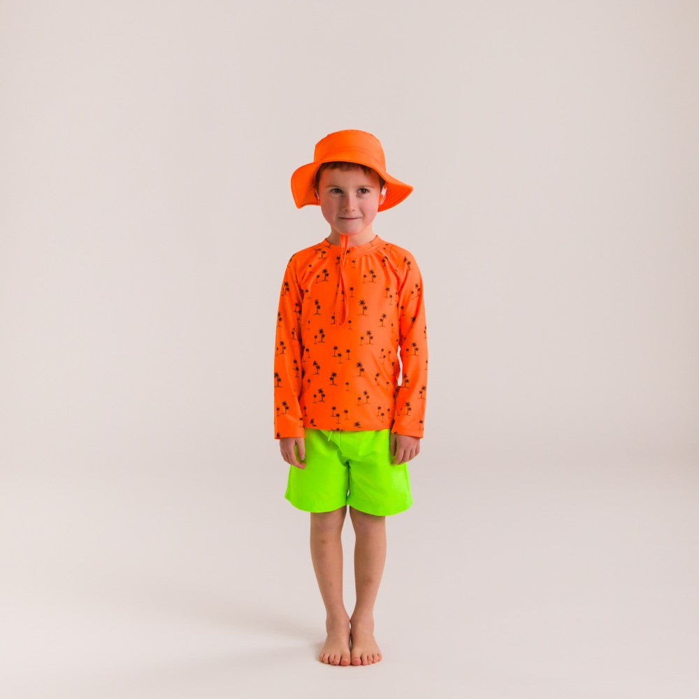 Child wearing an orange rash top, green shorts, and orange hat on a plain background