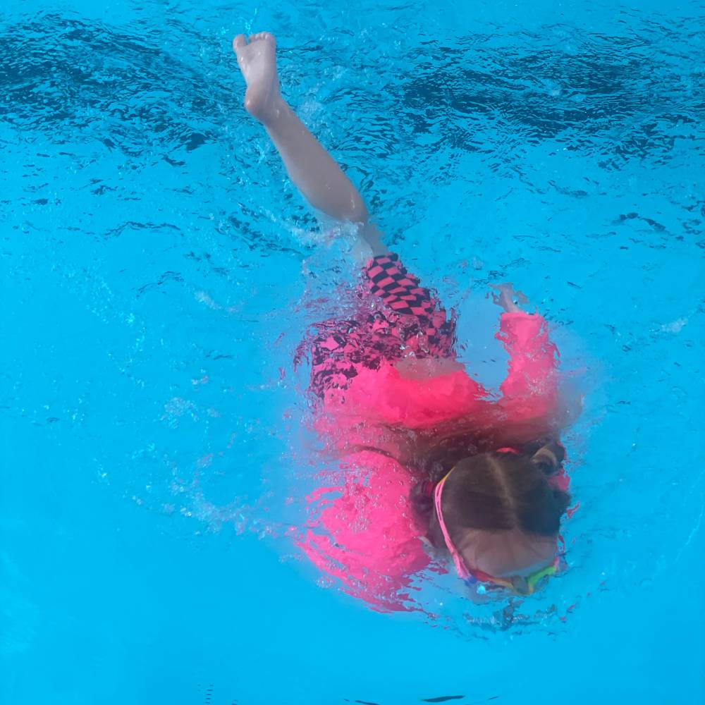 Child swimming underwater in a pool with neon pink rash vest and pink checkered swim shorts and goggles.