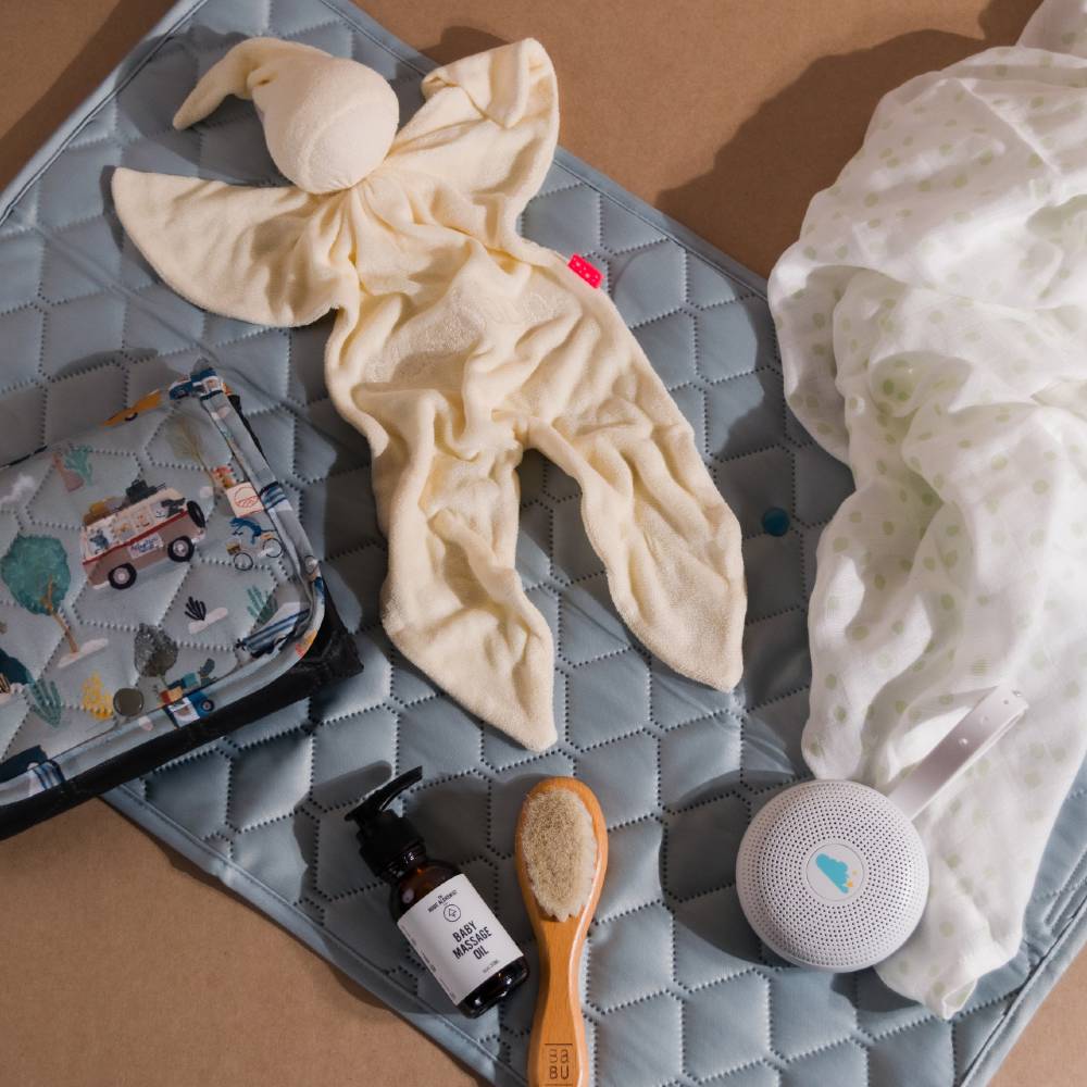 Baby care items on a nestling quilted change mat, including a swaddle, comforter and white noise machine with a folded change mat