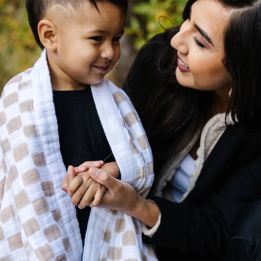 Woman holding a child's hand with a beige and white checkered baby blanket, surrounded by greenery