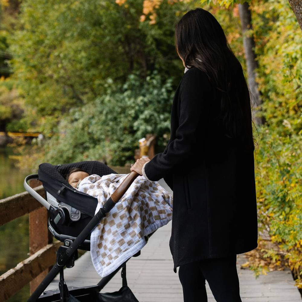 Woman pushing a stroller with a baby outdoors in a park with a beige and white checkered blanket in stroller