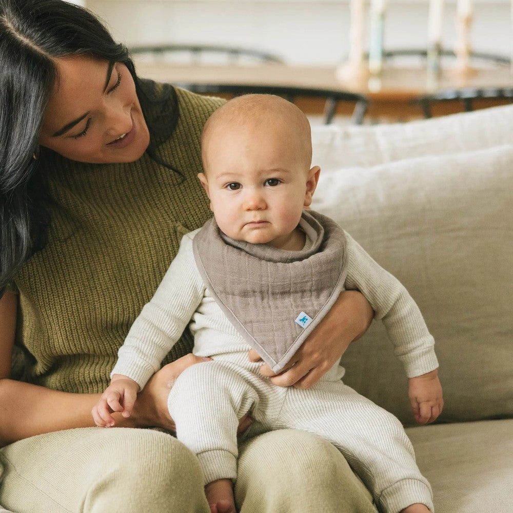 Woman holding a baby wearing a grey muslin bandana bib sitting on a couch