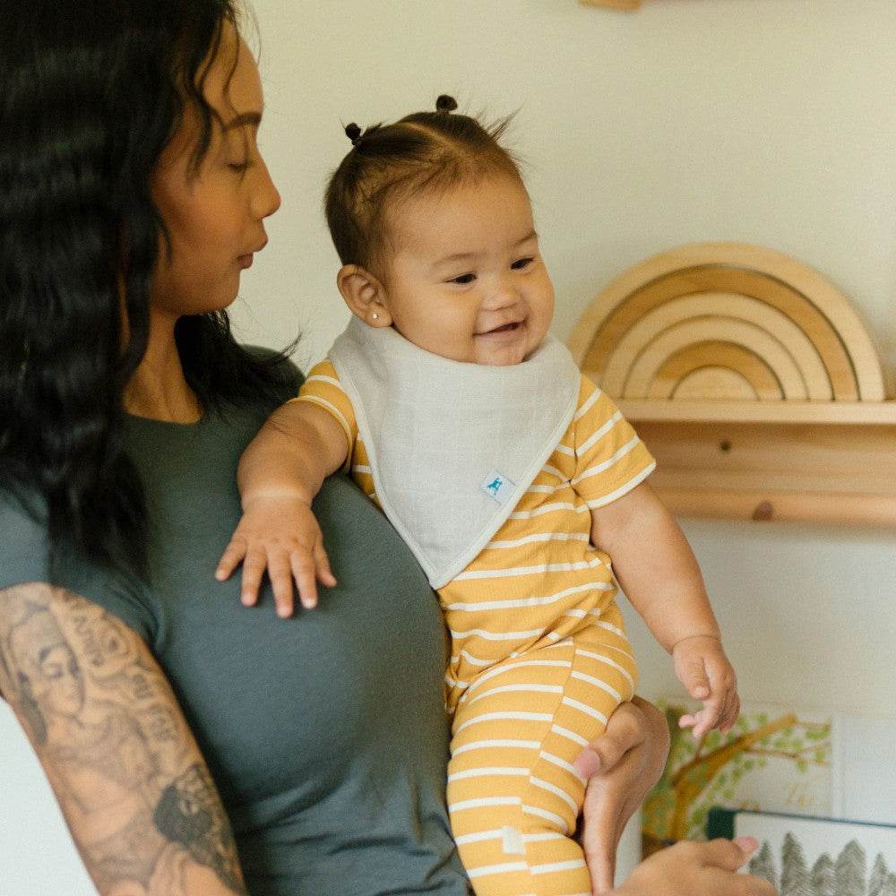 Woman holding a baby wearing a yellow striped outfit with grey muslin bandana bib , in a room with wooden shelves.